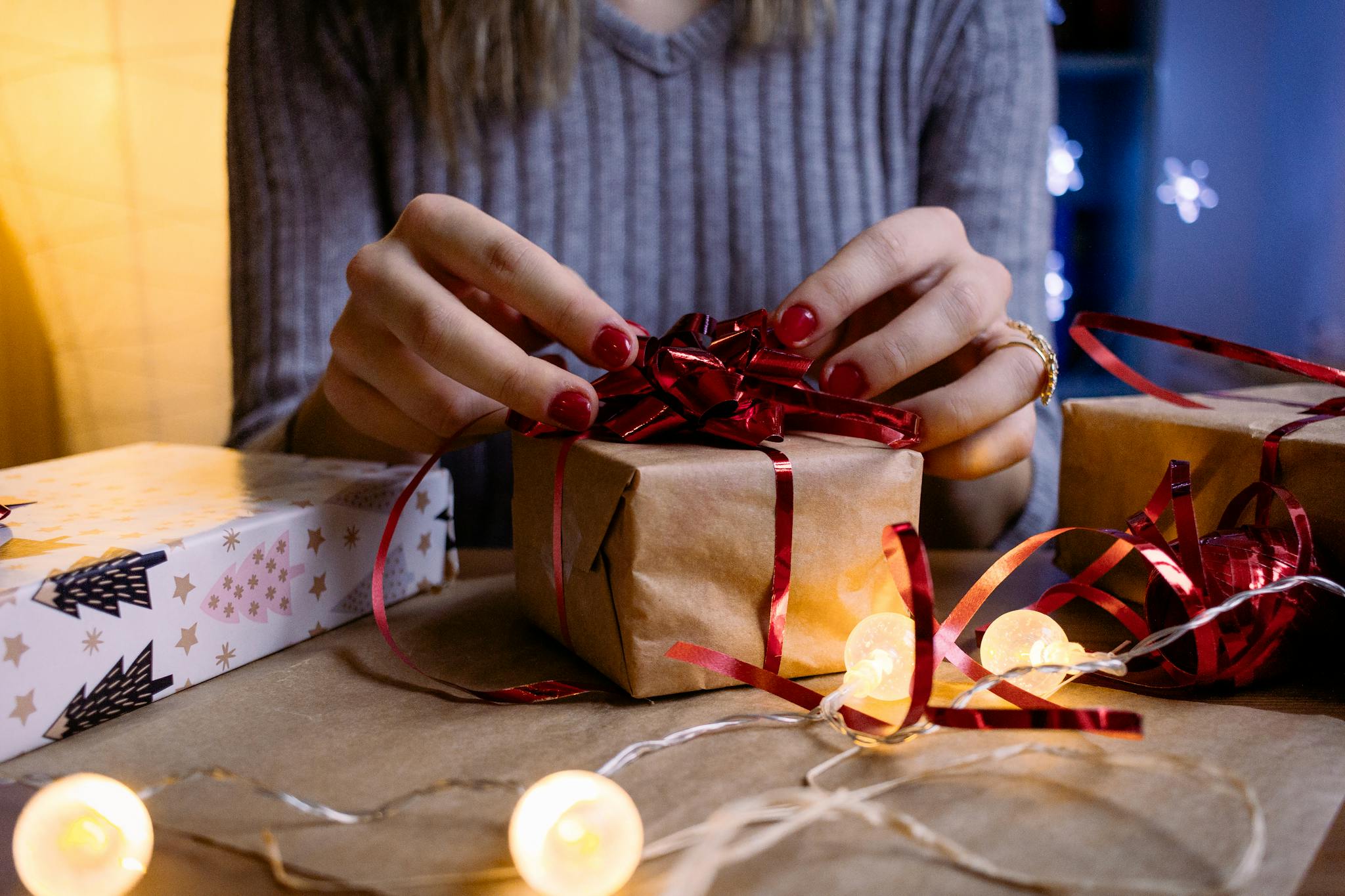Hands wrapping Christmas gifts with red ribbon amidst warm festive lights.